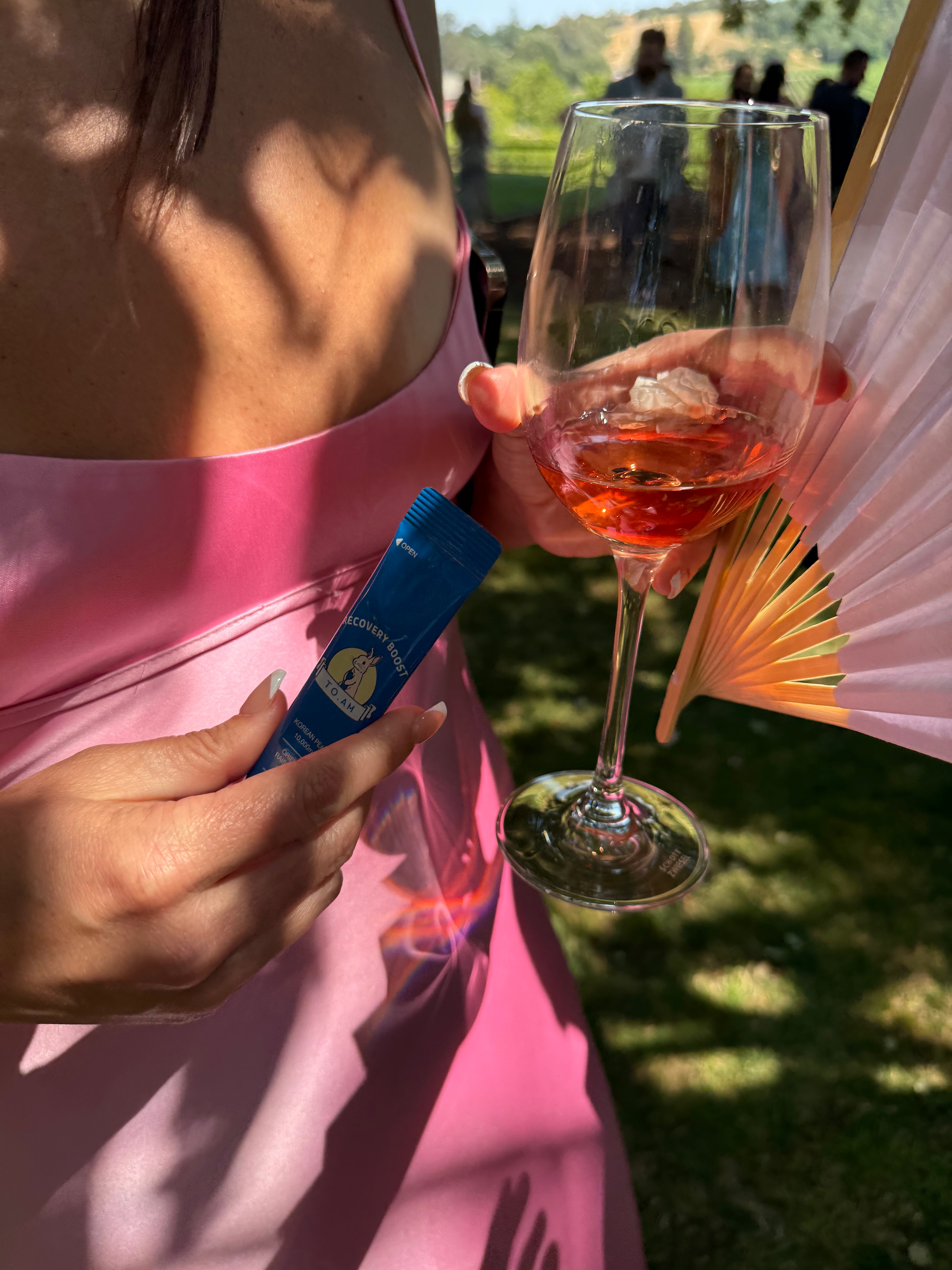 Person in a pink dress holding a glass of rosé wine and a TO. AM jelly stick, with a blurred outdoor background.